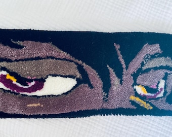 May include: A rectangular rug with a design of two stylized eyes. The eyes are in shades of purple, brown, white, and yellow against a black background. The rug has a textured, tufted appearance.