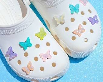 May include: White clogs decorated with colorful butterfly charms. The butterflies are in shades of blue, green, yellow, and purple. The clogs are on a light blue, glittery surface.