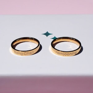 Könnte beinhalten: Zwei goldfarbene Ringe mit den Namen "Bang Chan" und "namjoonning" eingraviert.