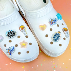 May include: White clogs with colorful Jibbitz charms. The charms include a star, a heart, a microphone, and a speech bubble that says "BOOM BOOM BASS!"
