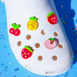 Puede incluir: Zapato blanco adornado con abalorios con forma de fruta. Los abalorios incluyen una fresa roja, una piña amarilla, un par de cerezas rojas y un melocotón rosa. Los abalorios tienen detalles verdes y están adheridos al zapato.