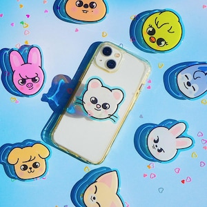 以下が含まれることがあります： 透明な携帯電話ケースに、さまざまな色の漫画の動物のステッカーが貼られています。ステッカーには、猫、ウサギ、ひよこ、犬、豚があります。携帯電話ケースは、白いハートが描かれた青い背景の上に置かれています。