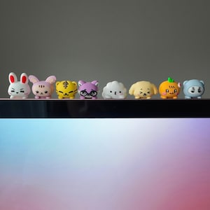 以下が含まれることがあります： カラフルな漫画風の動物フィギュアが8体並んでいます。白いウサギ、ピンクのウサギ、黄色のトラ、メガネをかけた紫色の猫、白い雲、黄色の犬、オレンジ色のカボチャ、青いクマが含まれています。