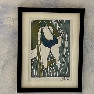May include: Framed print of a person in a swimsuit, viewed from behind, climbing a structure. The artwork is in shades of blue, green, and white, with a black frame and white matting. The print has a minimalist style.