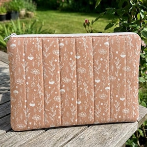 May include: A rectangular quilted laptop sleeve in a muted terracotta orange color featuring a white line-art botanical pattern of leaves and flowers. The sleeve has vertical stitching channels and a white zipper closure along the top edge.