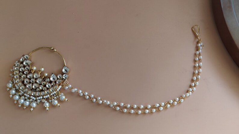 May include: A gold nose ring with a pearl chain. The nose ring is decorated with white stones and pearls.