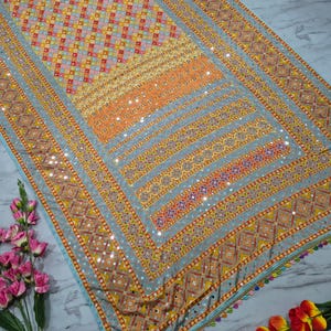 Op de afbeelding: Een gedetailleerd textiel met een geometrisch patroon in tinten oranje, geel en blauw. De stof heeft een rand met kleine spiegels en kleurrijke kwastjes. Het ontwerp is ingewikkeld en levendig, geschikt voor decoratieve doeleinden.