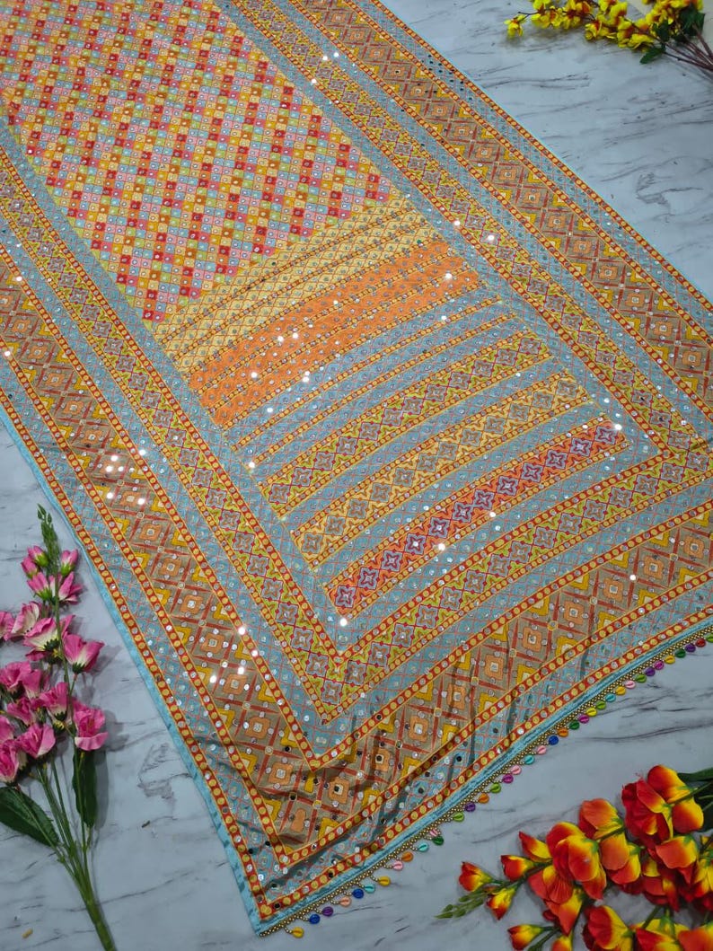 Op de afbeelding: Een gedetailleerd textiel met een geometrisch patroon in tinten oranje, geel en blauw. De stof heeft een rand met kleine spiegels en kleurrijke kwastjes. Het ontwerp is ingewikkeld en levendig, geschikt voor decoratieve doeleinden.