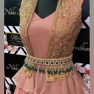 May include: A blush pink gown with a gold and green beaded belt. The gown has a layered skirt and a sheer, embroidered jacket.