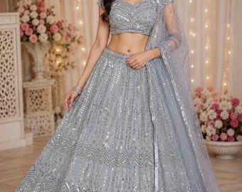 May include: A silver-gray, embellished lehenga choli set. The outfit features intricate sequin and beadwork, a cropped top, and a flowing skirt. A sheer dupatta drapes over the shoulder. The background includes floral arrangements and soft lighting.