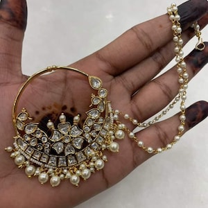 May include: A gold nose ring with a large, ornate design featuring white stones and pearls. The ring is attached to a chain with white pearls.