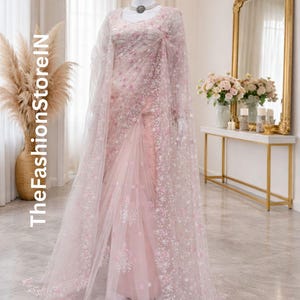May include: A pale pink saree with detailed floral embroidery and a sheer cape. The saree is displayed on a mannequin, highlighting its delicate design and flowing fabric. The FashionStoreIN text is visible on the left side of the image.