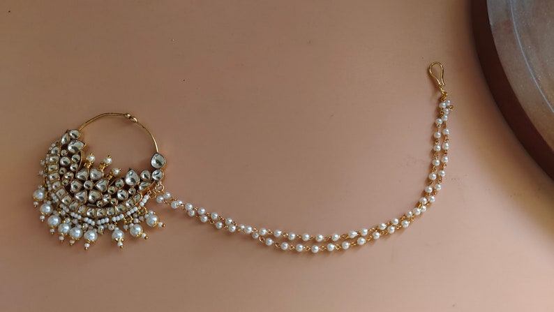May include: A gold nose ring with a large, ornate design featuring white pearls and clear stones. A delicate chain with white pearls hangs from the ring.