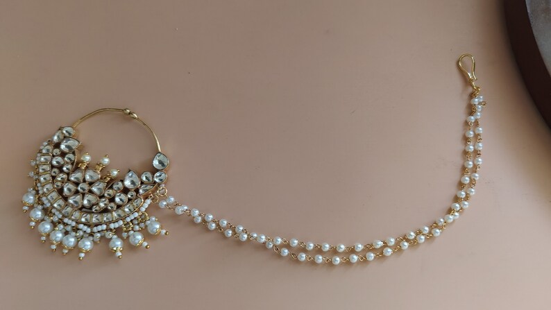 May include: A gold nose ring with a pearl chain. The nose ring is decorated with white stones and pearls. The chain is made of gold and pearls.