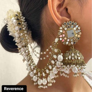 May include: Gold-toned earrings with a floral design and dangling pearls. The earrings feature a central mirror surrounded by smaller mirrors and colorful accents. The design includes a chain that attaches to a hair accessory with pearls and white flowers. The text "Reverence Picture" is in the bottom left corner.