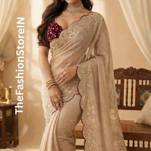 May include: A beige sari with gold embroidery and a scalloped edge. The blouse is maroon with gold detailing. The image includes the text "TheFashionStorelN".