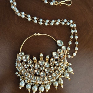 May include: A gold nose ring with a large, ornate design featuring clear stones and pearls. The ring is attached to a chain of pearls.