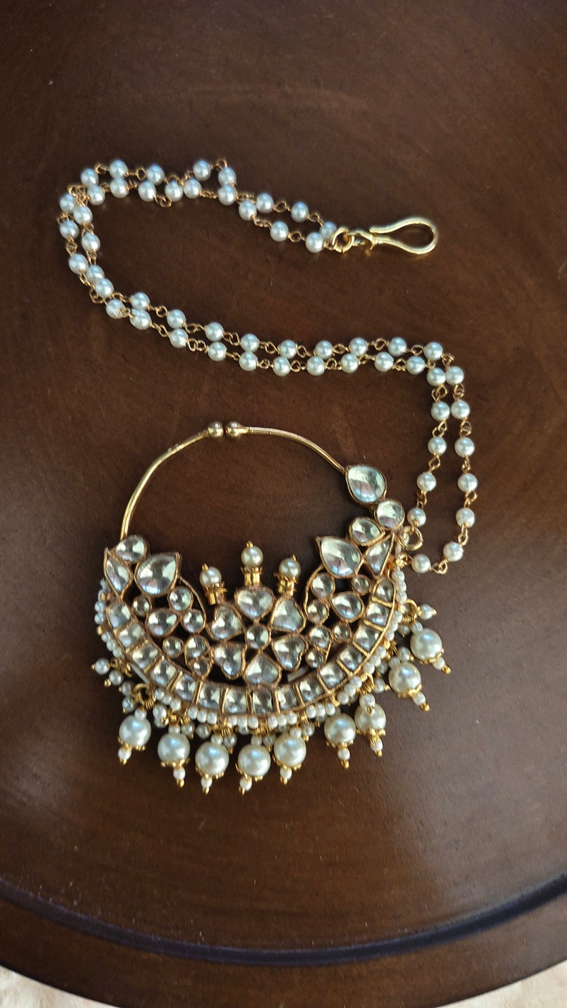 May include: A gold nose ring with a large, ornate design featuring clear stones and pearls. The ring is attached to a chain of pearls.