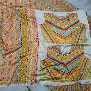 Op de afbeelding: Een kleurrijke textielset met een patroonontwerp met geometrische vormen en strepen in tinten geel, oranje, blauw en groen. De set bevat een lange lap stof en twee tops met een V-hals ontwerp, allemaal versierd met kleine, reflecterende versieringen.