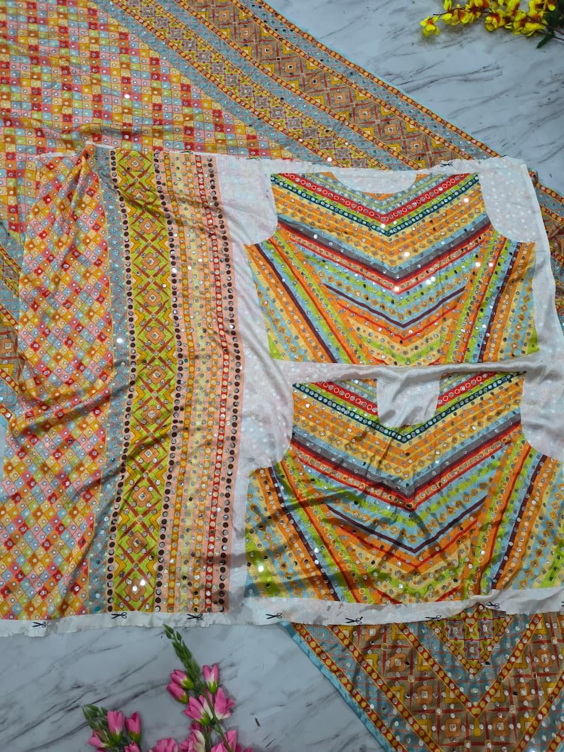 Op de afbeelding: Een kleurrijke textielset met een patroonontwerp met geometrische vormen en strepen in tinten geel, oranje, blauw en groen. De set bevat een lange lap stof en twee tops met een V-hals ontwerp, allemaal versierd met kleine, reflecterende versieringen.