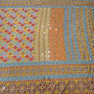 Op de afbeelding: Een kleurrijk textiel met een patchwork-ontwerp. De stof heeft een verscheidenheid aan patronen, waaronder ruiten, strepen en geometrische vormen, in tinten oranje, geel, blauw en rood. Kleine, reflecterende versieringen zijn over het ontwerp verspreid.