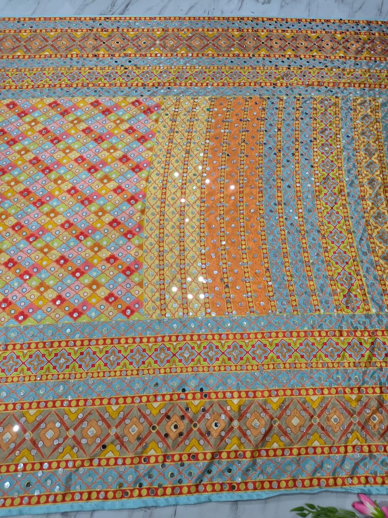 Op de afbeelding: Een kleurrijk textiel met een patchwork-ontwerp. De stof heeft een verscheidenheid aan patronen, waaronder ruiten, strepen en geometrische vormen, in tinten oranje, geel, blauw en rood. Kleine, reflecterende versieringen zijn over het ontwerp verspreid.