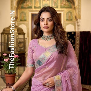 May include: A woman wearing a light purple sari with intricate embroidered borders and sleeves. The sari is paired with a matching blouse and a beaded choker necklace. The text "TheFashionStoreIN" is visible on the left side of the image.