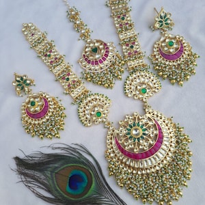 Op de afbeelding: Een sierlijke, goudkleurige sieradenset met oorbellen, hoofdbanden en een ketting. De stukken hebben een bloemen- en geometrisch ontwerp met groene en rode accenten en zijn versierd met kleine witte kralen. Een pauwenveer ligt in de buurt.