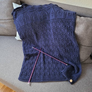 May include: A navy blue knitted blanket with a geometric pattern. The blanket is folded and laid on a gray couch with two knitting needles on top.