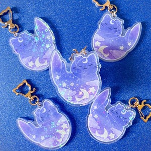 May include: Five iridescent purple and blue cat keychains with gold metal rings. Each keychain features a sleeping cat with a crescent moon on its head, surrounded by stars and clouds.