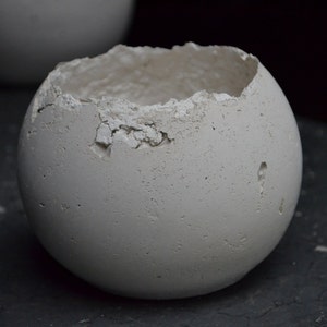 May include: A white, round, concrete planter with a rough, textured surface and a chipped opening at the top. The planter is sitting on a dark gray surface.