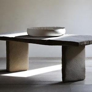 Puede incluir: Una mesa de piedra rústica con un cuenco de cerámica blanca en la parte superior. La mesa tiene una superficie rugosa y desigual y dos patas gruesas y rectangulares.