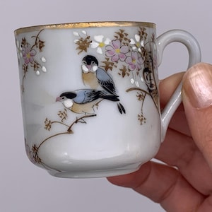 May include: Small white porcelain teacup with a gold rim and handle. The cup is decorated with a painted design of two birds perched on a branch with pink and white flowers. The teacup is held by a person.
