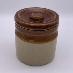 May include: A two-tone ceramic jar with a lid. The jar is cylindrical with a light beige base and a brown upper section and lid. The lid has a small knob on top. The jar is likely for kitchen storage.