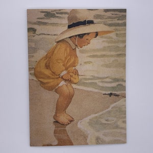May include: A young child wearing a yellow dress and a white sun hat is standing on a sandy beach, looking down at the waves.