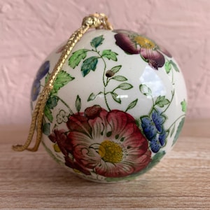 May include: A spherical white ceramic ornament decorated with a colorful floral pattern featuring large red and purple blossoms, green leaves, and small blue flowers. A gold-colored braided cord is attached to the top for hanging purposes.