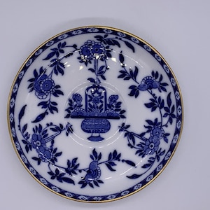 May include: A white porcelain saucer with a blue floral design and a gold rim. The centre features a blue floral basket. The rim is decorated with blue floral patterns and a gold border. A classic piece of tableware.
