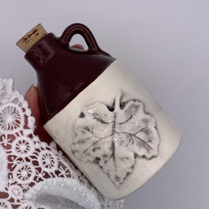 May include: A small ceramic jug with a dark brown glazed neck and a cream-coloured body. The jug features a raised, embossed leaf design on the front and is sealed with a light brown cork stopper. The surface has a smooth, glossy finish.