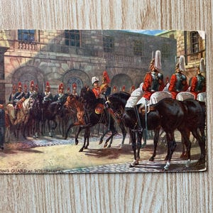 Op de afbeelding: Vintage ansichtkaart met de Mounting Guard in Whitehall. De afbeelding toont soldaten in rode jassen en hoge witte hoeden te paard, met een stenen gebouw op de achtergrond. De kaart heeft een licht verouderd uiterlijk.