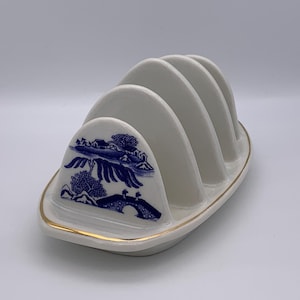May include: A white ceramic toast rack with four slots. The rack has a blue and white landscape design on the front and a gold trim around the base. The design includes a bridge, trees, and buildings.