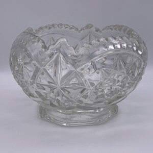 May include: A clear, cut-glass bowl with a scalloped rim and a starburst pattern. The bowl sits on a hexagonal base. The glass has a diamond pattern and is likely used for serving.