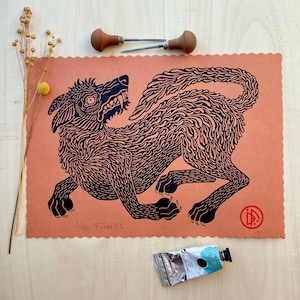 Peut inclure: Gravure sur linoléum en noir et blanc d'un loup grognant sur un fond de couleur pêche. L'impression est numérotée 1/100 et signée "Freeda '23".
