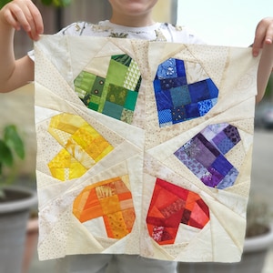 Peut inclure: Une courtepointe en tissu blanc avec un motif de patchwork composé de six cœurs arc-en-ciel. Les cœurs sont disposés en forme de trèfle.