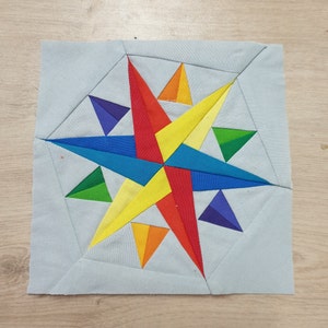 May include: A colorful patchwork quilt block featuring a star design with alternating triangles in shades of red, orange, yellow, green, blue, and purple.