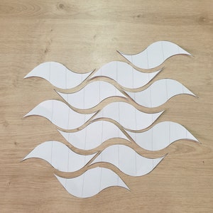 May include: A set of white paper shapes cut in the form of waves. The shapes are arranged in a wave pattern on a light brown wooden surface.
