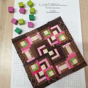 Peut inclure: Un bloc de patchwork Log Cabin fait à la main, composé de carrés de tissu marron, rose et vert. Le bloc est posé sur un papier blanc avec le texte "Logcabin quilt blocks". Plusieurs petits blocs de bois colorés sont éparpillés.