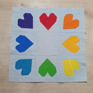 May include: A light blue fabric square with nine colorful felt hearts sewn on it. The hearts are arranged in a grid pattern with three rows and three columns. The colors of the hearts are red, orange, yellow, green, blue, purple, and a lighter shade of blue.