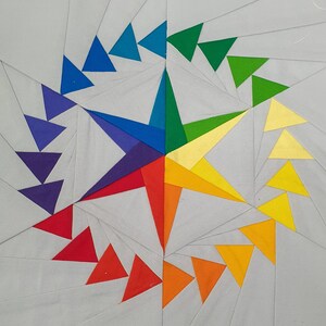 Puede incluir: Un bloque de colcha en forma de estrella de colores con un patrón en espiral. El bloque está hecho de tela en tonos de azul, verde, amarillo, naranja y rojo.