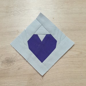 May include: A purple heart-shaped fabric patch is sewn into the center of a larger, light blue diamond-shaped fabric patch.