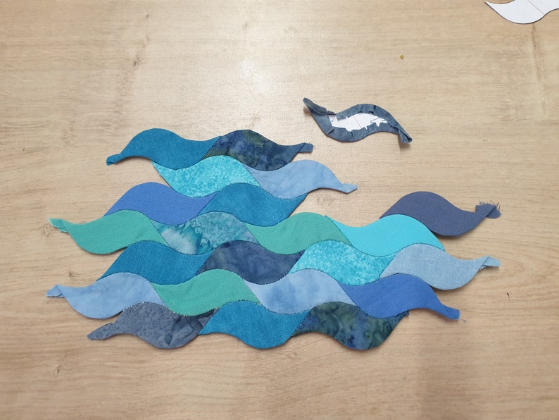 May include: A patchwork of blue, teal, and green fabric pieces sewn together in a wavy pattern, resembling ocean waves. The fabric pieces are arranged in a staggered pattern, creating a dynamic and textured look.